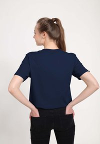 Námořnická modrá trička s krátkým rukávem a kulatým výstřihem, s hladkou texturou, v kombinaci s černými kalhotami. Model je vidět zezadu.