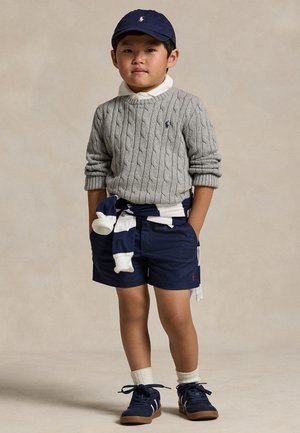 Giovane ragazzo in piedi con le mani in tasca, indossa un berretto blu navy, un maglione grigio a trecce, pantaloncini blu navy e sneakers blu navy con calzini bianchi.