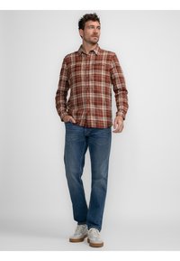 Camisa de cuadros abotonada en tonos apagados de rojo, marrón y crema, combinada con jeans azules y zapatillas claras. Presenta un cuello clásico y un bolsillo delantero.