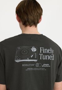 Camiseta gris oscuro con un estampado blanco de un tocadiscos y el texto "Finely Tuned", además de detalles de branding adicionales. Diseño básico de cuello redondo.