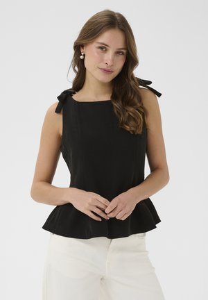 Jonge vrouw met lang golvend haar, gekleed in een mouwloze zwarte top met gestrikte schouderbandjes en een witte broek, staand tegen een effen achtergrond.