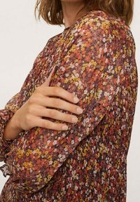 Blusa de estampado floral con mangas largas y fruncidas. Presenta una mezcla de flores rojas, amarillas y blancas sobre un fondo oscuro, fabricada con un tejido suave y transparente.