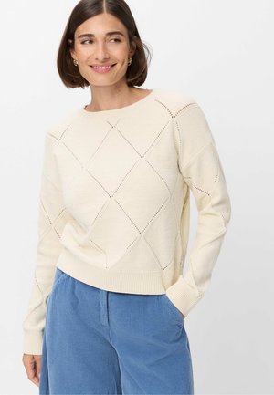 Donna con capelli corti castani che indossa un maglione crema a maglia con motivi a diamante e pantaloni di velluto a coste blu, sorridente e che guarda di lato.