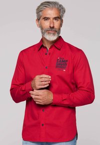 Rotes Langarmhemd mit Knöpfen am Kragen, mit einem marineblauen Logo, dunklen Knöpfen und einem geraden Saum, getragen über einer hellblauen Jeans.