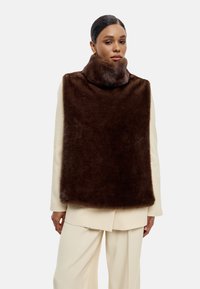 Hnědý umělý kožich s vysokým límcem, který má jemnou strukturu a obdélníkový tvar, nošený přes krémový outfit.