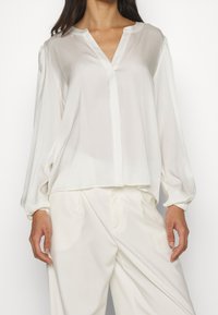 Weiße, durchsichtige Bluse mit langen Ärmeln, V-Ausschnitt und lockerer Passform. Sie hat geraffte Ärmel und eine glatte Textur mit minimalen Nähdetails.