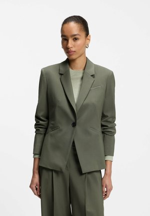Mujer con chaqueta y pantalones de traje verde oliva combinados con una camiseta verde clara, con el cabello recogido y pequeños pendientes de aro.