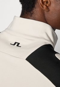 Collar sweatshirt i lys beige og svart, med teksturert stoff og en svart logo på baksiden av nakken. Rene linjer og moderne design.