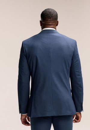 Man draagt een nauwsluitend donkerblauw pak en staat met zijn rug naar de kijker op een effen beige achtergrond.