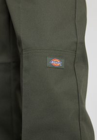 Tamsiai žalia medžiaga su tekstūruotu paviršiumi. Ant šono siuvinėta maža "Dickies" logotipo etikete, vaizduojanti spalvingą emblemą.