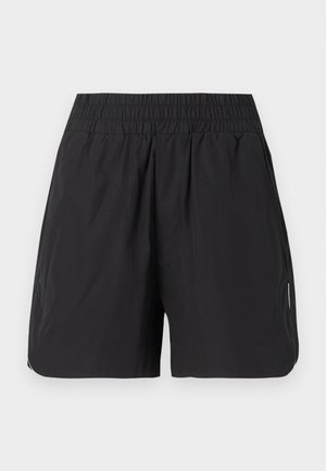 Γυναίκα όρθια με το χέρι στη μέση, φορά λευκό crop top, μαύρο σορτς, λευκές κάλτσες και μπεζ-μαύρα αθλητικά παπούτσια σε λευκό φόντο.