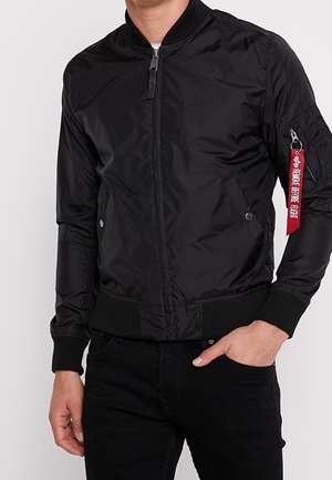 Blouson noir en nylon brillant, avec fermeture éclair, poignets côtelés et poches latérales, agrémenté d'une étiquette rouge sur la manche.