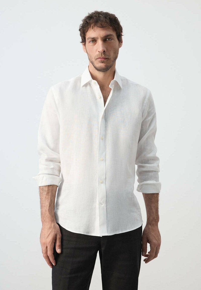 Homme debout devant un fond uni, portant une chemise blanche à manches longues retroussées et un pantalon noir.
