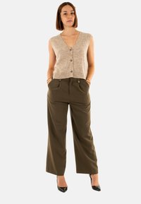 Gilet senza maniche in maglia beige con bottoni, abbinato a pantaloni larghi verde oliva. Il modello indossa tacchi a punta neri.