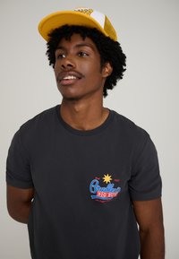 Dark gray cotton t-shirt featuring a colorful "Bradley's Big Buy" logo with a star design. Complemented by a yellow and white trucker cap.