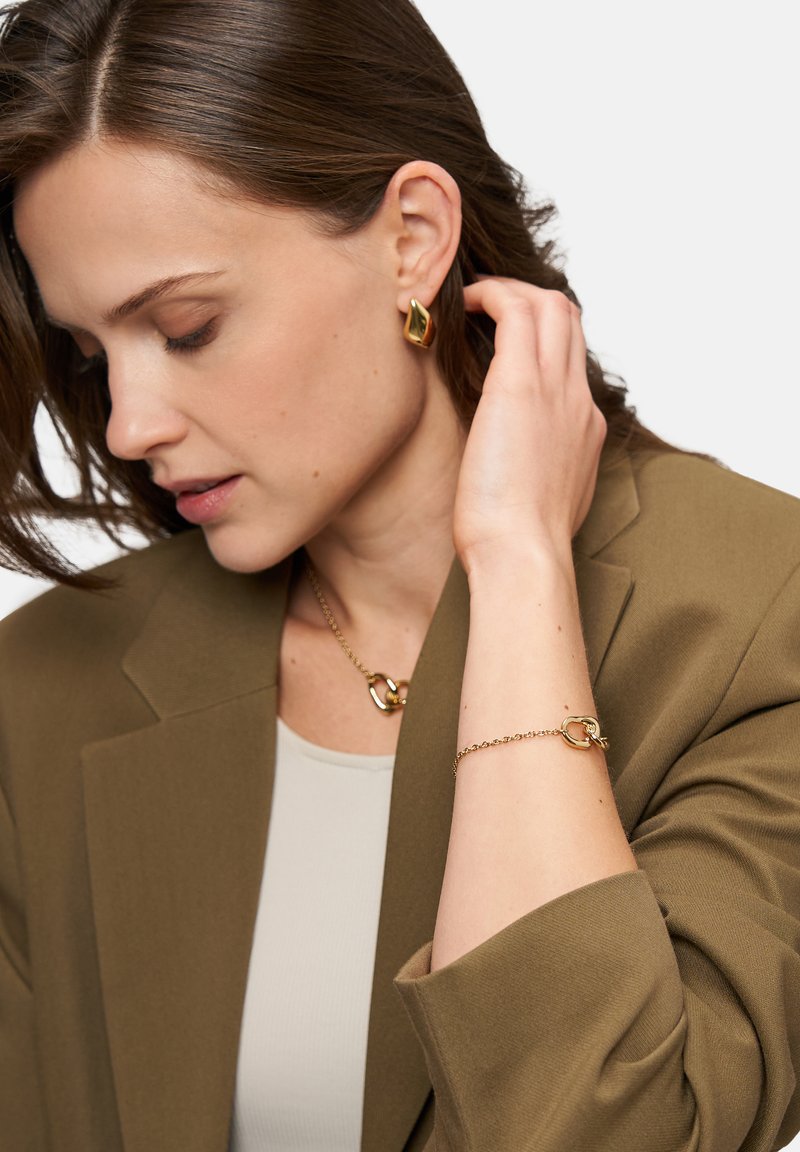 Gold jewelry including earrings, bracelet, and necklace; smooth, shiny, modern design; worn with a light top and brown blazer.