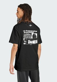 Black cotton t-shirt featuring a graphic print on the back with “superstar PARIS” text, and Adidas logo. Short sleeves, crew neck design.
