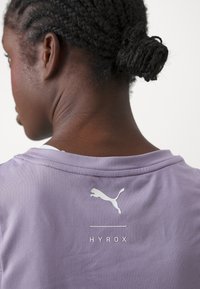 Ljubičasta atletska majica s bijelim Puma logotipom i tekstom "HYROX" na leđima. Tkanina izgleda glatko, s okruglim izrezom.