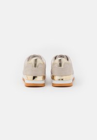 Beżowe zamszowe trampki z metalicznym złotym akcentem na pięcie, białą podeszwą i teksturowaną, brązową gumową podeszwą zewnętrzną. Zaokrąglony tył z przeszyciami.