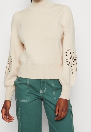 Pull à col roulé crème à manches longues avec motifs floraux découpés sur les manches, porté avec un pantalon cargo bleu sarcelle avec des coutures blanches.