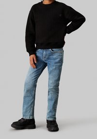 Niño vestido con un suéter negro acolchado, jeans azul claro y botas negras, de pie con una mano en la cadera contra un fondo gris.