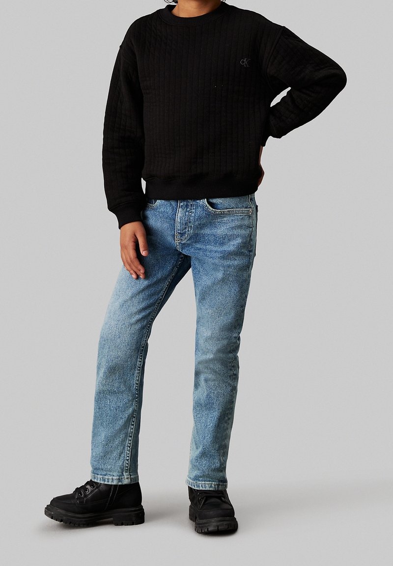 Niño vestido con un suéter negro acolchado, jeans azul claro y botas negras, de pie con una mano en la cadera contra un fondo gris.