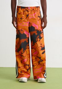 Person iført løse orange bukser med abstrakte rosa og sorte mønstre, hvide sneakers, stående på grøn gulv mod en hvid væg.