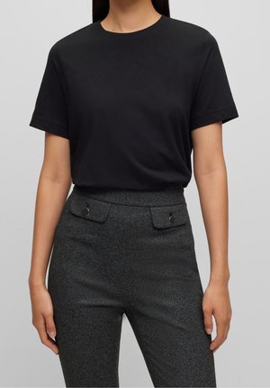 Femme portant un t-shirt noir uni à manches courtes, rentré dans un pantalon taille haute gris foncé texturé avec des poches à rabat et des boutons.