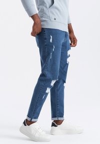 Hombre vestido con un sudadera gris clara, jeans azules desgastados arremangados en los puños y zapatillas blancas con toques negros en el talón, de pie de lado.