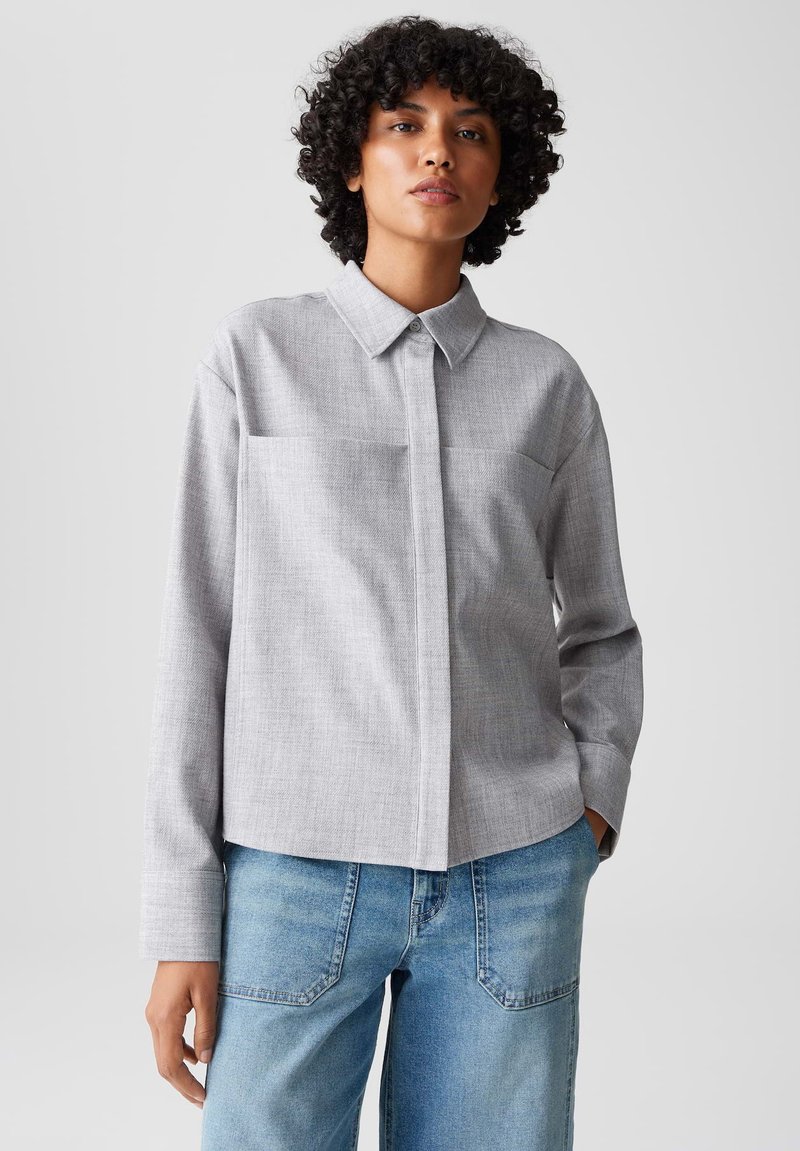Persona con capelli ricci che indossa una camicia collare grigia chiara e jeans blu, in piedi contro uno sfondo chiaro semplice.
