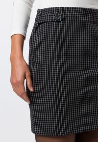 Femme portant une jupe à carreaux noir et blanc avec un détail de bouton, un haut à manches longues blanc et des collants noirs transparents, la main reposant sur le côté.