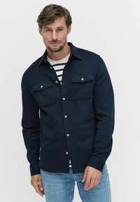 Mörkblå skjorta med knappar, två bröstfickor, tillverkad av textilier med struktur, bärs ihop med en randig skjorta under, tillsammans med ljusblå jeans.