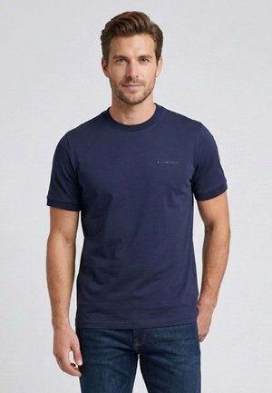 Uomo con capelli castani corti che indossa una maglietta a maniche corte blu navy e jeans blu scuro, in piedi davanti a uno sfondo chiaro.