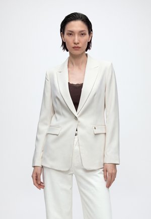 Femme aux cheveux courts et foncés portant un blazer blanc sur un haut marron foncé et un pantalon blanc, debout devant un fond clair uni.
