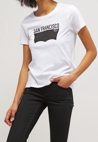 Persona vistiendo una camiseta blanca con el texto y logo en negro de "San Francisco", combinada con pantalones negros, con la mano reposando en el bolsillo.