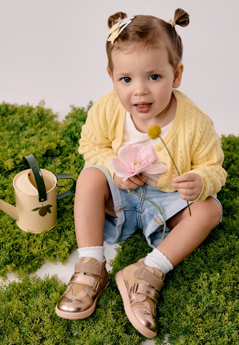 Tout-petit avec deux petites couettes portant un cardigan jaune et des chaussures dorées, assis sur de la mousse verte, tenant des fleurs roses et jaunes à côté d'un arrosoir.