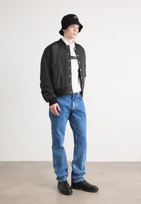 Giacca bomber nera sopra una camicia bianca, jeans blu e stivali neri. Logo rosso chiaro e bianco sul cappellino. Vestibilità morbida e casual in tutto.