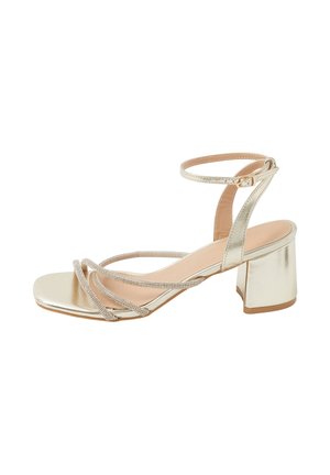 Sandalo con tacco a blocco metallizzato dorato, con sottili cinturini incrociati, chiusura con fibbia alla caviglia e design con punta quadrata.