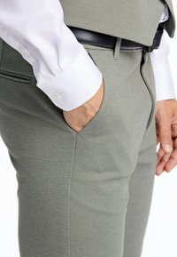 Graue Tailored-Hosen mit schrägen Taschen, strukturiertem Stoff und schwarzem Ledergürtel. Weißer Hemdärmel am Handgelenk sichtbar.