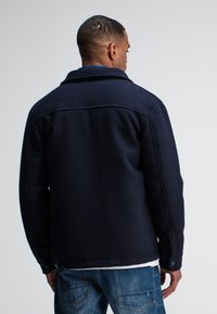 Navy wool-blend jacket with a structured collar, buttoned cuffs, and back yoke detail. Paired with faded blue denim jeans.