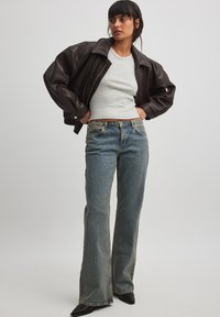 Chaqueta de cuero marrón, camiseta gris corta y jeans de campana azul claro. El modelo está de pie con zapatos de tacón negros frente a un fondo neutral.