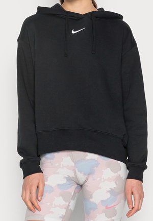 Osoba ubrana w czarną bluzę z kapturem marki Nike z troczkami oraz pastelowe legginsy w chmurki z różowym logo Nike na lewym udzie.