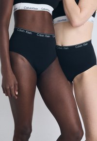 Mutandine alte nere con logo di Calvin Klein, tessuto liscio. Due modelle posano vicine, mettendo in risalto i toni di pelle e gli stili contrastanti.