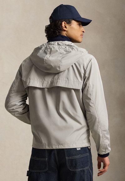 Φως γκρι μπουφάν με κουκούλα, κατασκευασμένο από ελαφρύ υλικό, με ενίσχυση στην πλάτη και ριμπ μανσέτες. Συνδυασμένο με σκουρόχρωμο τζιν παντελόνι.