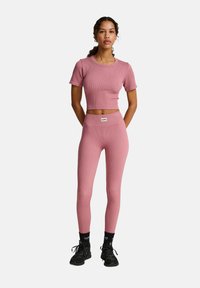 Roze geribde crop top en legging set, met een getailleerd ontwerp, zachte textuur en een klein labeldetail op de tailleband. Zwarte schoenen en sokken.