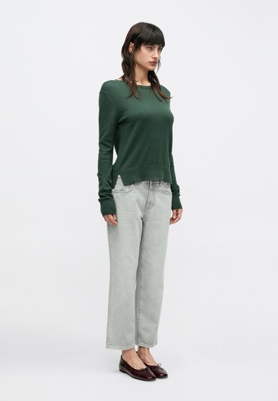 Σκούρο πράσινο πλεκτό πουλόβερ με μακριά μανίκια και ριμπ μανσέτες, συνδυασμένο με ανοιχτό γκρι, φαρδιά τζιν παντελόνια και σκούρα καφέ φλατ παπούτσια. Απλό φόντο.