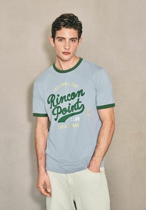 Ung mand med kort krøllet hår iført en lyseblå Rincon Point surfklub t-shirt og lyse bukser mod en ensfarvet baggrund.
