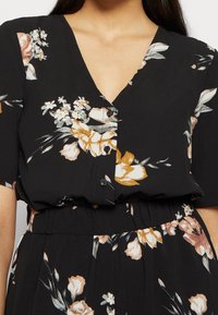 Vestido negro con flores que presenta un escote en V, mangas cortas y una cinturilla fruncida. Adornado con patrones florales en beige y rosa.