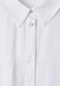 Chemise blanche à col avec une patte de boutonnage avant comportant deux boutons visibles, un en métal et un en plastique, sur un tissu légèrement texturé.