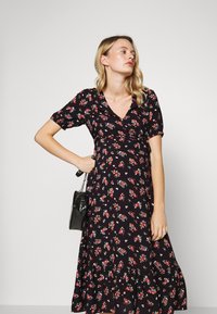 Vestido preto floral com mangas curtas, apresentando um decote em V e uma cintura franzida. A saia tem uma barra com babados e pequenas flores vermelhas e brancas.